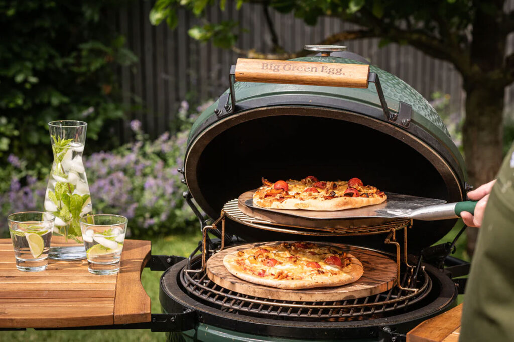 Big Green Egg grill with pizzas cooking on ceramic plates in a backyard setting