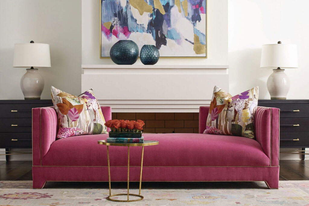 A channel-tufted sofa in deep pink velvet styled with colorful floral throw pillows and a patterned area rug — a refined example of pattern drenching using a solid anchor piece with expressive accents