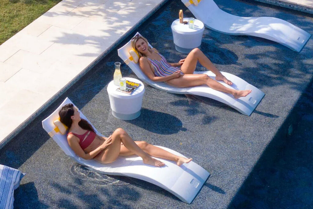 An overhead view of two women relaxing on sleek, white Ledge Lounger chairs positioned inside the shallow tanning ledge of a luxury swimming pool with white accent tables.